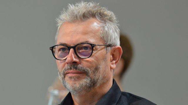 Portrait von Markus Jung. Ein Mann mittleren Alters mit grau melierten, kurzen Haaren. Er trägt ein schwarzes Hemd und eine Brille mit dunklen Brillengestell. Sein Gesichtsausdruck ist neutral bis leicht nachdenklich. Der Hintergrund ist unscharf und grau.