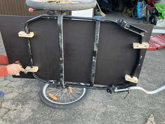 Ein aufgekippter Fahrradanhänger. Mensch sieht von unten dagegen. Es sind die Holzklötze zu sehen, mit denen die Siebdruckplatte am Rahmen befestigt wurde.