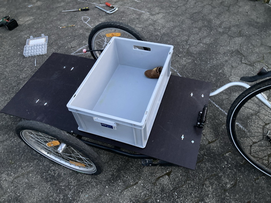 Fahrradanhänger von oben am Rad angekuppelt. Das Hauptteil besteht aus einer Siebdruckplatte. Oben auf steht eine Eurobox zum Größenvergleich, die eher klein wirkt.