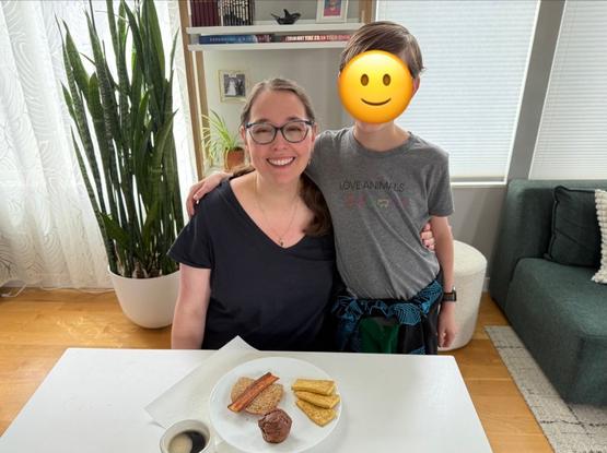 Andrle, a smiling white woman with glasses, sits at the end of a white dining table in front of a plate of brunch. Next to her stands a boy with his arm around her shoulder. His face is obscured with a smiling emoji. In the background is a tall snake plant and a bookshelf with some knick-knacks. The shades on the windows behind are closed.