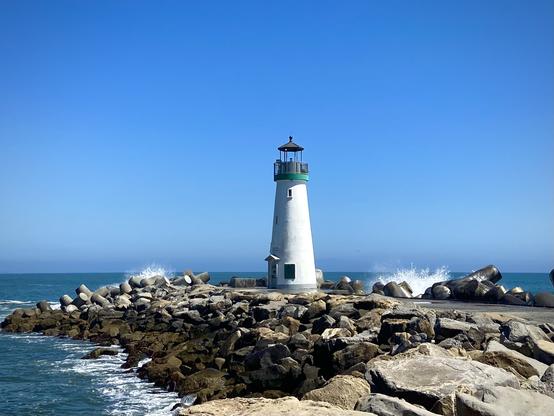 A white lighthouse with a green top stands at the end of a rocky jetty, extending into the ocean. Waves crash against the large rocks surrounding the lighthouse. The sky is clear and blue, creating a bright and sunny atmosphere. The lighthouse has a glass lantern room at the top, and the scene captures the beauty of the coastline and the crashing waves.