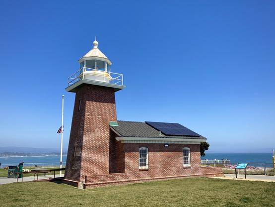A brick lighthouse building stands on a grassy area under a clear blue sky. The lighthouse has a white, dome-shaped top with a glass lantern room. Solar panels are visible on the roof of the building. Nearby, a flagpole holds a small American flag at half-mast. In the distance, the ocean and coastline are visible. There are a few people in the background, some near informational signs. The scene is bright and sunny, suggesting a pleasant day at the coast.