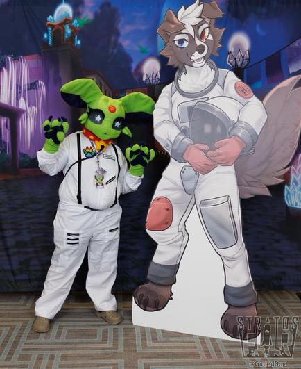 A photo of Cosmo, an alien Espeon fursuit, next to a standee of Laika, the Stratosfur mascot. A watermark in the corner indicates that the photo was taken by Gilded Dog.