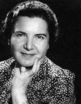 A portrait photo of the face and shoulders of a mature woman with a blouse in spots. She holds two fingers by her cheek.