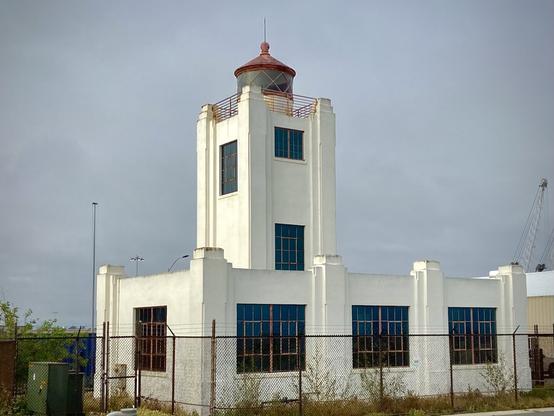 A white, art moderne style lighthouse building stands against a cloudy sky. The structure has a red roof with a small lantern room at the top. Large, blue-framed windows cover the sides of the building, and a chain-link fence surrounds the property. Nearby, utility equipment and a crane are visible in the background, giving the area an industrial feel. The lighthouse is somewhat isolated, with sparse vegetation at the base.