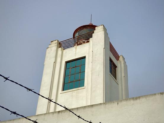 A close-up view of the top of a white lighthouse building. The lighthouse has a red roof and a small lantern room with a railing around it. A large blue window is visible on the side of the structure. In the foreground, a barbed wire fence runs along the bottom of the image, adding an industrial feel to the scene. The sky is mostly overcast and grey, and the building shows some weathering.