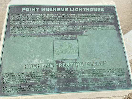 A bronze plaque detailing the history of the Point Hueneme Lighthouse. The plaque is titled 'POINT HUENEME LIGHTHOUSE' and provides historical information about the lighthouse’s construction, its evolution over time, and its original Fresnel lens. The plaque also mentions the history of the name 'Hueneme,' derived from the Chumash word for 'resting place.' There is a small map or illustration at the center of the plaque showing the lighthouse's structure.