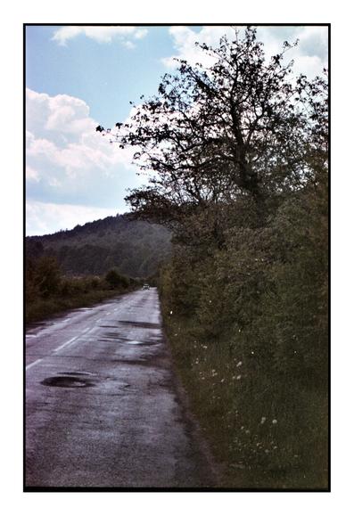 A slightly faded film photo of a rural road bordered by trees and hills, with a single car in the distance under a partly cloudy sky. The colors are muted, and there are signs of film grain and age artifacts, giving the image a nostalgic, vintage atmosphere.