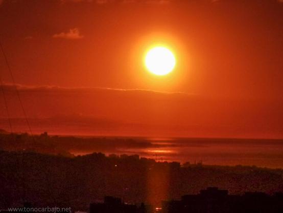 Salida de sol sobre Barcelona. Abajo la línea de ciudad a contraluz. En la zona media el reflejo sobre el mar, una tenue línea de nube y en la parte media superior el sol. Toda la foto está teñida por su luz cálida y anaranjada.