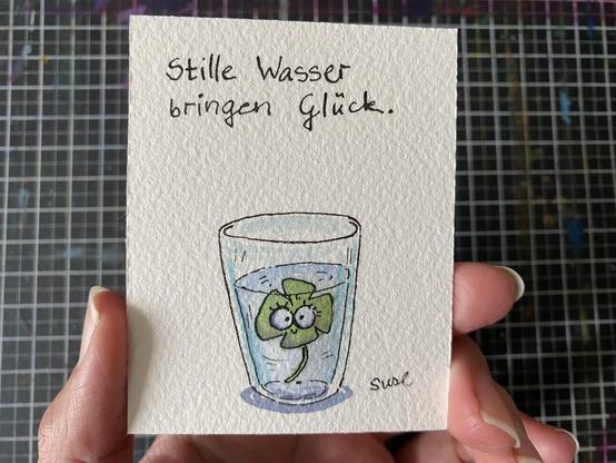 "Stille Wasser bringen Glück." Ein Glas Wasser, in dem ein vierblättriges Kleeblatt schwimmt. Es lächelt freundlich.