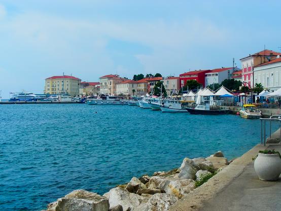 This image captures a serene waterfront scene. In the foreground, there's a rocky shoreline with a concrete walkway. The water is calm and blue, reflecting the clear sky above. Along the waterfront, several boats are docked, including fishing boats and a larger vessel. The background features a row of colorful buildings with red, yellow, and white facades, adding a vibrant touch to the scene. Some buildings have outdoor seating areas with white umbrellas, suggesting cafes or restaurants. The overall atmosphere is peaceful and inviting, with a blend of natural beauty and charming architecture, making it a picturesque coastal setting.