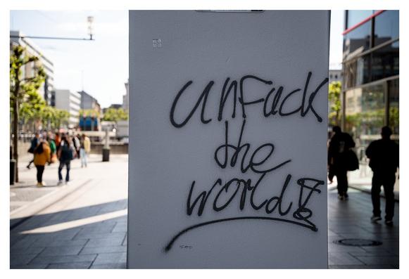 Ein grauer Mauerstein wirkt als Träger für einen Schriftzug in schwarzer Sprühfarbe, der die Worte „UNF*CK the WORLD“ zeigt. Im Hintergrund sind Menschen zu sehen, die sich in einer urbanen Umgebung bewegen, umgeben von modernen Gebäuden und angepflanzten Bäumen. Das Wetter ist klar und sonnig, was die Szene lebendig und dynamisch erscheinen lässt.