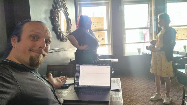 Dan gestures at his laptop in a quiet pub function room, on which he's writing some code. In the background, two women are having a conversation.