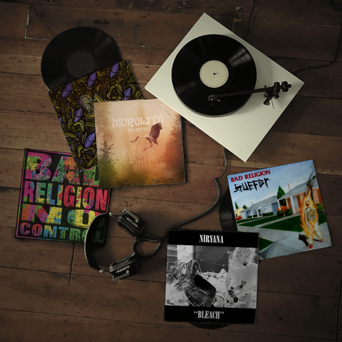 Das Bild zeigt eine Sammlung von Vinyl-Platten und einer Plattenspielerplatte auf einem Holzboden. Im Zentrum befindet sich ein moderner weißer Plattenspieler mit einer schwarzen Plattenspielerplatte, auf der ein Tonarm ruht. Um den Plattenspieler herum liegen mehrere Vinyl-Albumcover. Links oben ist ein Albumcover mit einem farbenfrohen, floralen Muster zu sehen. Direkt daneben ist das Albumcover von "Nonolith Dystopia" mit einem Bild eines Storchs in einem Dämmerlichtszenario. Unten links ist das Albumcover von "Bad Religion In a Control" mit einem bunten, psychedelischen Design. Rechts daneben ist das Albumcover von "Bad Religion Suffer" mit einem Cartoon-ähnlichen Bild eines brennenden Menschen vor einem amerikanischen Suburban-Haus. Unten in der Mitte ist das Albumcover von "Nirvana Bleach" mit einem Schwarz-Weiß-Bild zu sehen. Neben den Albumcover liegt ein Paar schwarzer Kopfhörer.