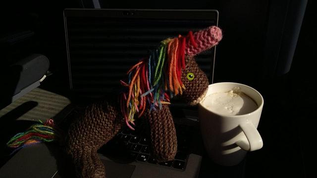 Max, das bunte Strickhorn, mit einem ICE-Cappuccino in der Sonne auf dem Laptop auf dem Klapptisch.