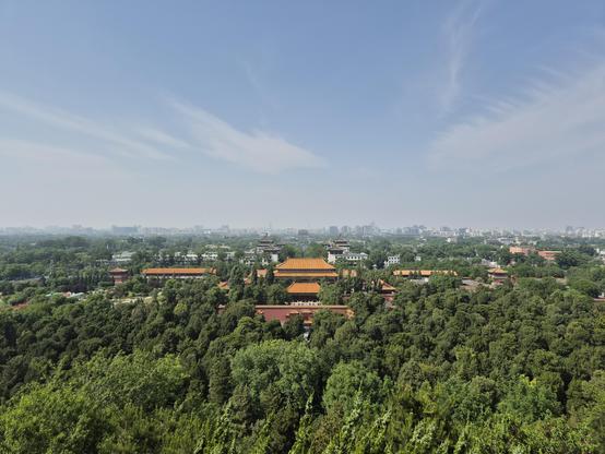 Panoramic view from a hill towards the northern part of Beijing, green trees in the foreground with buildings in traditional Chinese architecture and more urban city skyline further in the distance.