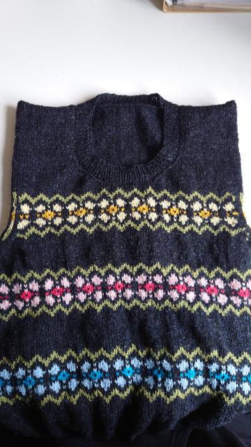 Shows a black vest top (tank top or sweater vest) with three horizontal striped flower style motifs. The bottom is blue, the centre is pink, and the top is yellow. The neck is a high crew neck with ribbing. The sleeves are still knitted together. It's lying in a white table lit by daylight.
