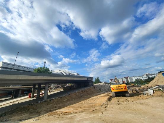 Großbaustelle vor freundlich bewölktem blauen Sonnenhimmel. Links ein Stück Autobahnbrückenauffahrt, die abgebrochen ist. Dahinter das Dach des ICC Berlin mit eine Spitze Berliner Funkturm. Rechts ein gelber Bagger von hinten an großem Sandhaufen. In der Ferne Wohnhäuser, die sonst an der alten Autobahn angrenzten.