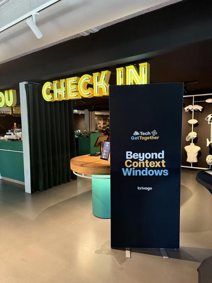 An interior scene featuring a welcoming area with a prominent yellow neon "CHECK IN" sign. In the foreground, a black banner displays the text "Beyond Context Windows" alongside the Trivago logo. There is a circular wooden table with informational materials.