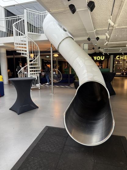 An indoor space features a large, shiny tube slide extending from a spiral staircase. Nearby, there is a tall, black cocktail table and several people are visible in the background. The setting appears modern and inviting, with colorful decor.