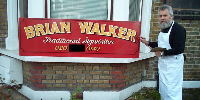 Man posing with sign painter's tools in front of a hand-painted sign resting on a window ledge. The sign is on a red background and the lettering is headed up by the name 'Brian Walker', under which is 'Traditional Signwriter' and a phone number.