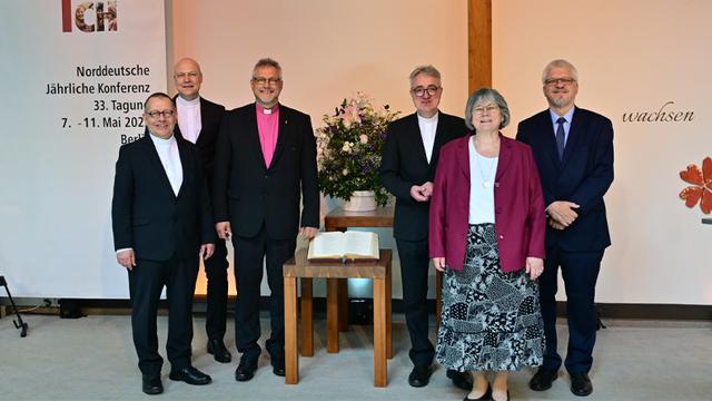 Hinter den sechs Personen, die sich zum Foto aufgestellt haben, steht links das Motto der Konferenztagung, »Auf dich vertrau ich«, mit weiteren Angaben zur Tagung, rechts ein großes Kreuz und in der Mitte auf zwei Tischen eine Bibel und ein Blumenstrauß.