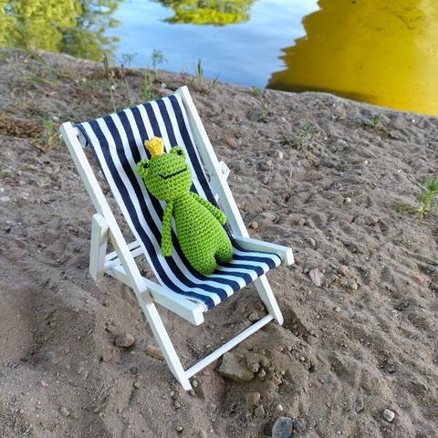 A picture of a crocheted amigurumi frog in a tiny doll's beach chair, on some sand, next to the water of a canal.