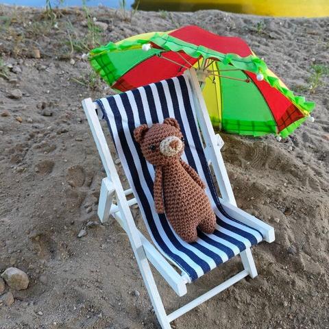 A picture of a crocheted amigurumi bear in a tiny doll's beach chair, on some sand, next to the water of a canal. There's a tiny parasol next to the chair.