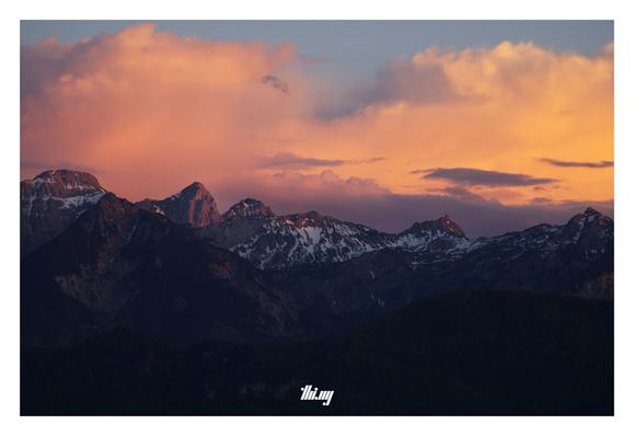 Photo of layers of mountain peaks & ridges of 3 valleys during sunset. Vanilla clouds/sky. The clouds with a rich orange/purple glow and only some of the peaks still catching the last (red) light. Lower parts already in deep shade, only silhouettes. Higher up parts still have some remnants of the last snow, but considerably less than last year...
