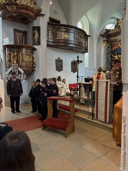 Das Innere einer katholischen Kirche. Der Feuerwehrkommandant steht neben mehreren Kindern der Feuerwehrjugend in Uniform. Dahinter sitzen zwei Ministranten auf einer Bank. Der Pfarrer steht daneben, am Sprechpult steht der Kommandant-Stellvertreter, ebenfalls in Uniform.