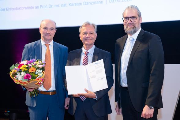 Three people in suits smile at the camera. The person on the left is holding a bouquet of flowers, the person in the middle is holding a certificate.