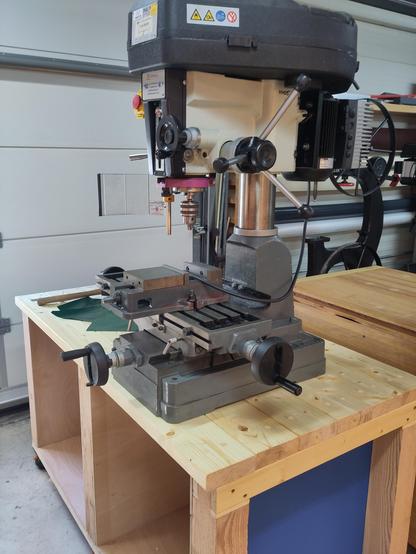 Photo of a table top mill standing on a woden table. The mill has an grey cast iron base where the X-Y table is placed up on. The top of the machine is resting on a bare metal cylinder that allows it to move up and down. The top of the machine is white and has an drill holder. The motor of the mill is behind the mill and the top of the mill is a plastic protection for the belt drive mechanism. The mill has both a drill press style quill feed handle (placed on the right side of the machine) and an fine adjustment quill handle that works via a worm-gear (placed on the normal quill feed and reachable via a wheel at the front of the machine, next to the depth gauge)