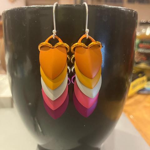 A pair of scalemail earrings hanging from a black coffee mug. Each earring consists of five overlapping scales, in the colours of one of the lesbian pride flags - dark orange, light orange, white, pink, violet.