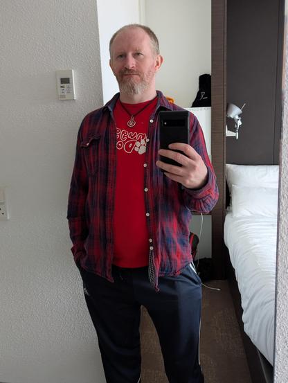 Human Dog, wearing a casual outfit of tracksuit pants, a t-shirt and a flannel check shirt. A necklace with round dog ID tag is clear to see as he's taking a photo in a mirror with his mobile phone.