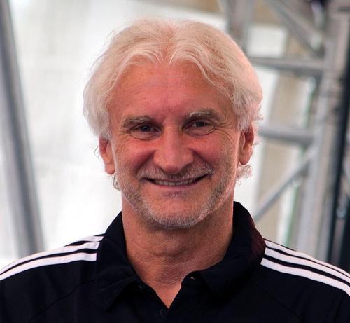 Foto von Rudi Völler. Er trägt ein schwarzes Sportoberteil mit weißen Streifen. Im Hintergrund sind verschwommene Strukturen zu sehen, die möglicherweise Teil einer Sporteinrichtung sind.