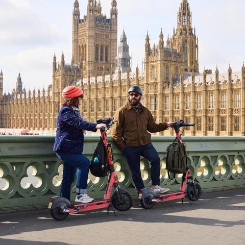 The image shows two people with electric scooters on a bridge, with the Houses of Parliament in London visible in the background. On the left, one person is wearing a bright orange helmet, a navy blue jacket with red spots, blue jeans, and white trainers, facing the other person. On the right, the second individual is wearing a black helmet, brown jacket, and dark trousers. Both are resting against their pink scooters. The bridge has a light green railing with intricate circular patterns. The grand architecture of the Houses of Parliament, with its detailed Gothic Revival style, towers, and spires, forms the backdrop under a partly cloudy sky.

TfL Image - Voi E-scooter