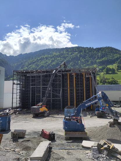Foto. Industrieareal mit Baumaschinen vor einer geöffneten Gebäudefassade.
