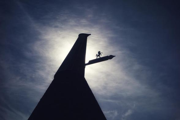 A low-angle, silhouetted view of a traditional oast house against a bright, cloudy sky. The conical roof of the oast house dominates the left side of the frame, leading up to a cowl. A weather vane, featuring a small horse figure, extends horizontally from the side of the cowl towards the right. The sun is positioned behind the oast house and clouds, creating a bright halo effect and silhouetting the structure and the weather vane. The sky transitions from a bright, almost white area behind the oast house to a darker, more textured blue in the upper and right portions of the image.