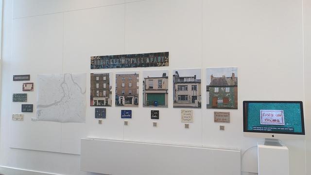 A white wall with an exhibition art project set up on it. On the left a column of 5 ceramic tiles and mosaic. Then an A0 map. Then 5 A2 pictures of derelict buildings each with a ceramic tile and a QR code under. On the right a computer screen displaying a video with interviews and shots of derelict buildings. On top of it all a big mosaic title written "Don't let me fall apart".