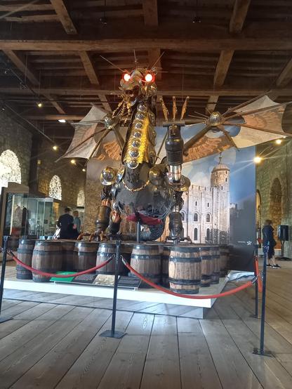 A huge scary dragon with glowing red eyes. It is made from various types of weapons and armour. It stands on a raft of gunpowder barrels in a stonewalled room with a wooden ceiling and floor.