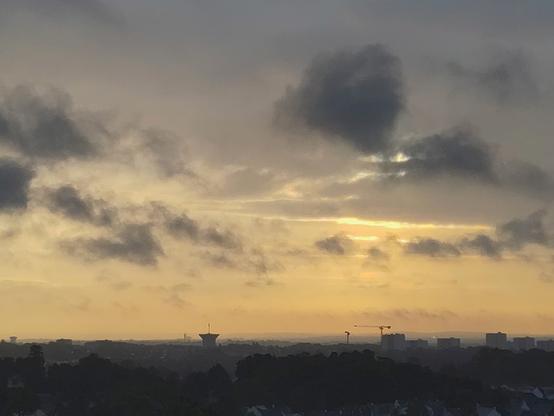 le ciel est jaune et gris, des nuages traversent l'horizon et plus bas des bâtiments s'élèvent du sol sombre, on voit leur forme se découper clairement notamment des immeubles, une grue et un château d'eau