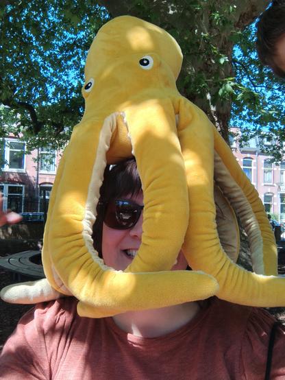 Een juf met zonnebril zit buiten bij de monumentale plataan verstopt onder een grote gele knuffel octopus op haar hoofd