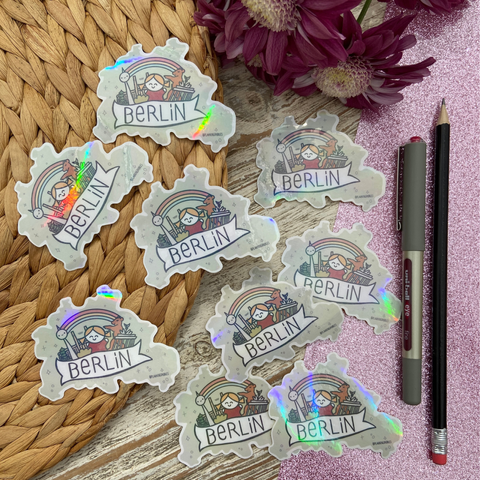 Mehrere meiner handgemachten Berlin-Sticker mit Fernsehturm, Olympiastadion, Brandenburger Tor, Berliner Bär, Regenbogen und meiner Signatur-Figur Pabby. Die Grundform ist die Silhouette der Stadt Berlin. Im Hintergrund zartlilafarbene Blumen.