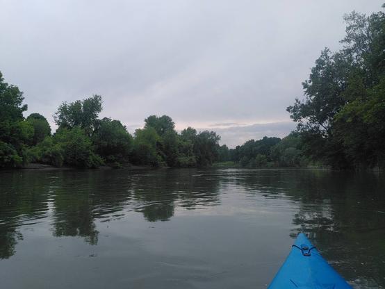 The bow of a blue kayak can be seen on a wide and calm river. The shores are lined with trees and the sky is overcast.