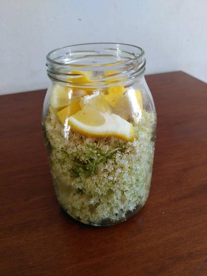 A glass jar filled with elderflower and lemon stands on a dark wooden table.