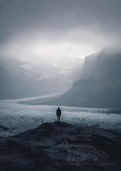 A person stands on a rocky ledge overlooking a vast glacier surrounded by foggy mountains. The scene is cold and dramatic, with muted tones and a sense of solitude. The image has a watermark in the bottom right corner that reads "Sandner Art & Photography" with a geometric fox logo.