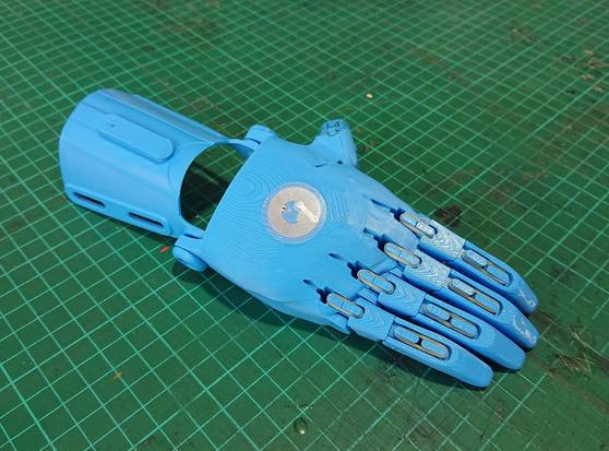 A 3D print of an E-nabling the Future open-source prosthetic hand, printed in blue plastic.  The small metal NFC sticker for Museum in a Box to recognise can be seen on the back of the hand.