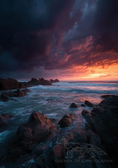 A dramatic seascape at sunset, featuring jagged rocks emerging from swirling ocean waves. The sky is split between deep indigo storm clouds on the left and a fiery orange-pink sunset on the right, casting vivid reflections across the water and wet rocks in the foreground. The mood is both turbulent and serene, capturing the raw beauty of nature’s contrasts.