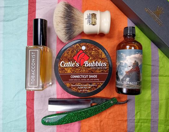 (Le Chat Mistral description)

The image showcases a collection of grooming items arranged on a striped cloth. At the centre, there's a tin of "Catie's Bubbles" shaving soap with a label indicating "Connecticut Shade" and scents of sweet tobacco, vanilla, oak, and nutmeg. To the left, there's a bottle of perfume labelled "Tobacconist." On the right, a bottle of aftershave named "Trail Tobacco" is visible. Above the soap, there's a shaving brush and a small container of shaving cream. Below the soap, a green-handled straight razor is placed. The items are neatly organised, suggesting a traditional shaving setup.