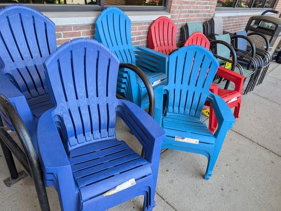 A photo of another set of bike racks outside a different building, where brightly-colored plastic lawn chairs are set up between the racks. Clearly bike racks are actually just reserving space for seasonal inventory.